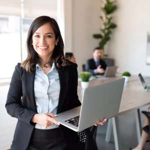 Portrait of smiling female entrepreneur holding a laptop with team in background at office conference room